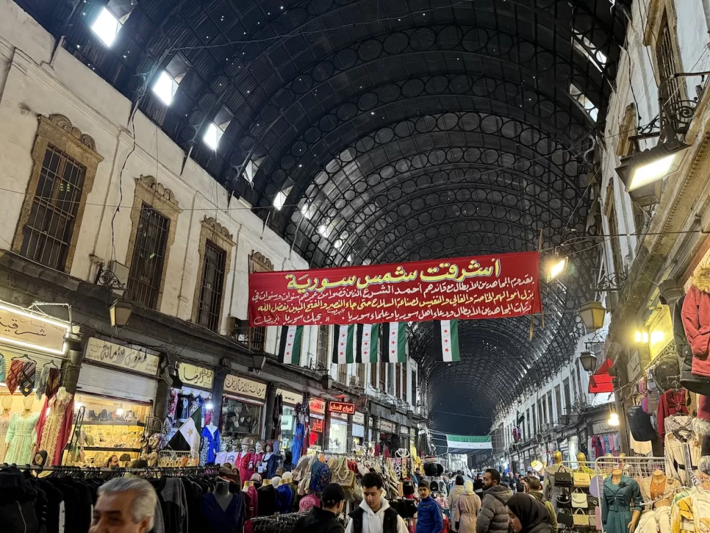 A bustling corridor of Al-Hamidiyah Souq is filled with shoppers, textiles, and souvenirs.